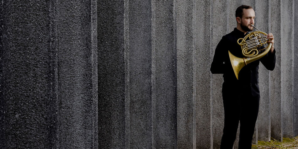 Photo of Gabriel Trottier playing the horn in front of a cement wall