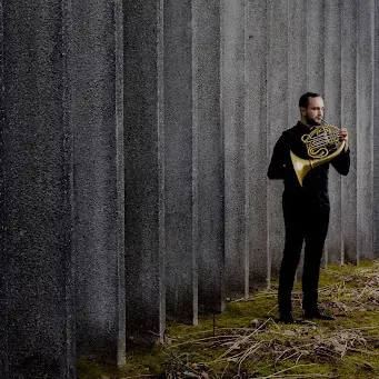 Photo of Gabriel Trottier playing the horn in front of a cement wall